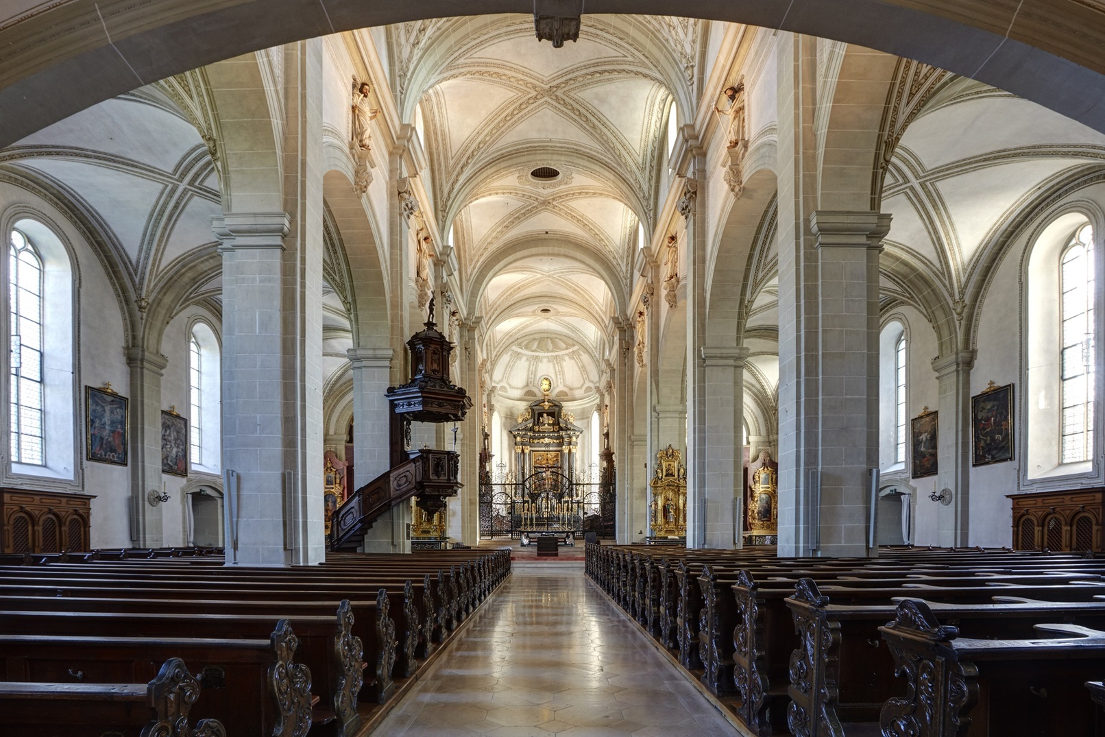 Blick durch das Mittelschiff der Hofkirche St. Leodegar in Luzern in Richtung Hochaltar. Beidseitig reihen sich dunkle Holzbänke, Pfeiler tragen das Kreuzgewölbe, links steht eine barocke Holzkanzel.