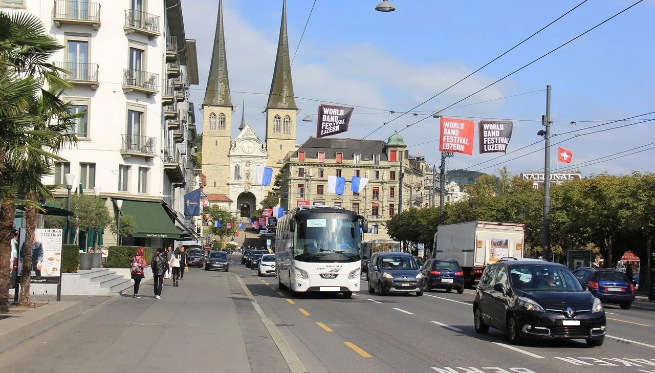 Weisser Reisecar auf einer belebten Stadtstrasse in Luzern, im Hintergrund die Türme der Hofkirche und Werbebanner des World Band Festival Luzern.