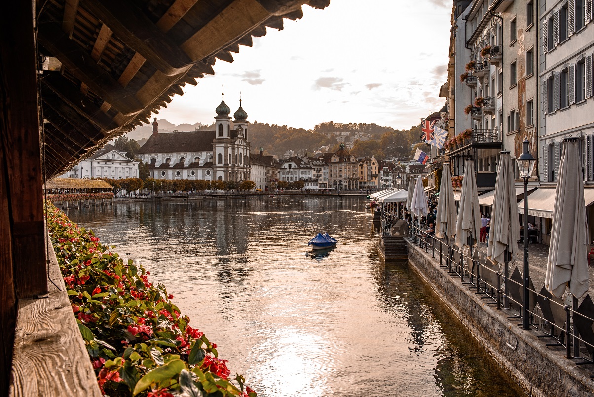 Blick von der überdachten Kapellbrücke in Luzern über die Reuss auf die weiss-barocke Jesuitenkirche im Hintergrund, flankiert von der historischen Altstadt und einem Restaurantufer mit Sonnenschirmen, im warmen Herbstlicht.
