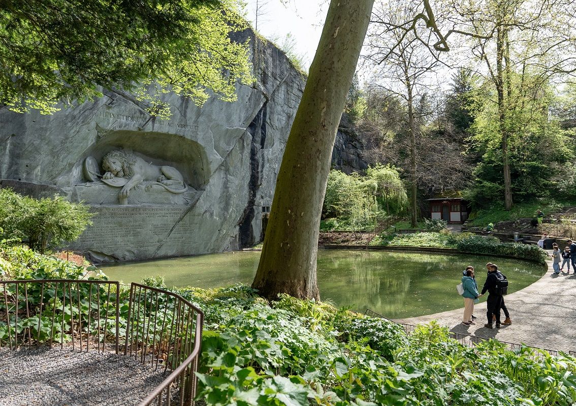 In einen Sandsteinfelsen gehauenes Relief eines Löwen, davor ein ovaler Teich, umgeben von Bäumen und Grünpflanzen. Mehrere Personen spazieren auf dem Weg entlang des Teiches.