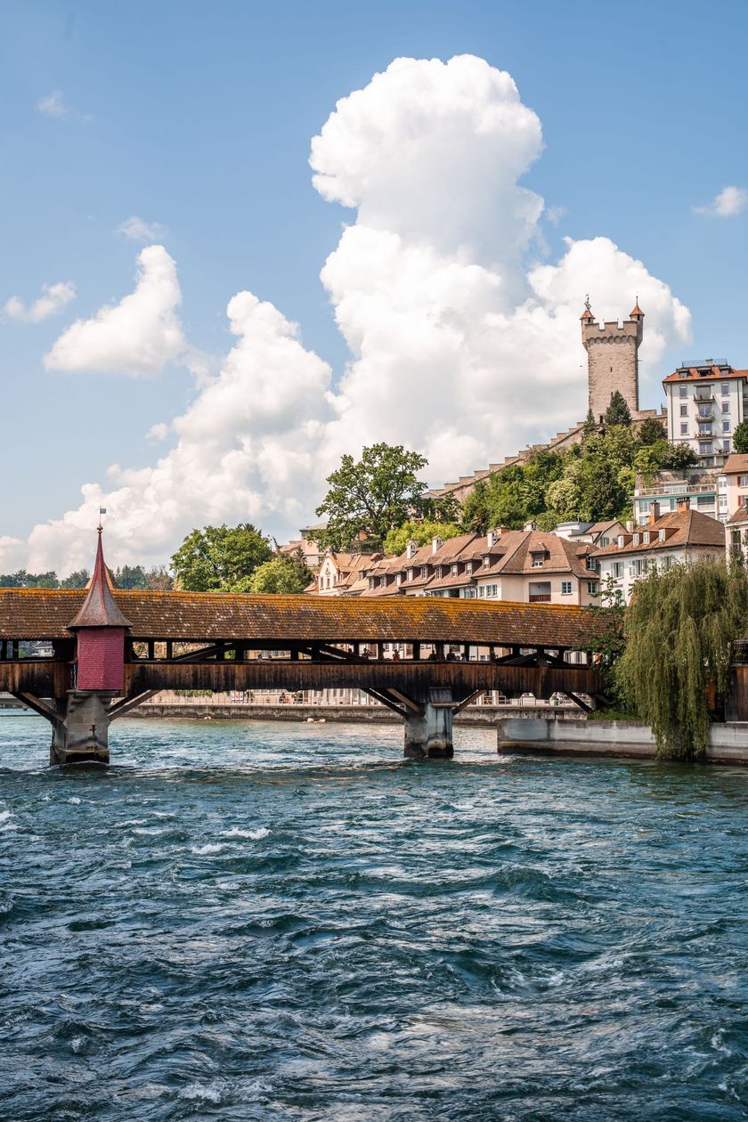 Die überdachte hölzerne Spreuerbrücke überspannt die Reuss in Luzern. Im Hintergrund sind Altstadthäuser mit Ziegeldächern sowie ein Turm der Museggmauer auf einer bewaldeten Anhöhe sichtbar. Der Himmel ist blau mit grossen Quellwolken.