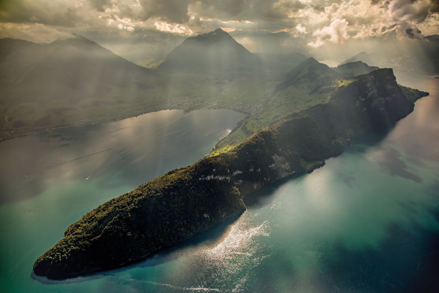Luftaufnahme des Vierwaldstättersees mit einer langgezogenen grünen Halbinsel, steilen Uferhängen und Bergen im Hintergrund, durchzogen von warmem Sonnenlicht und dramatischen Wolken.