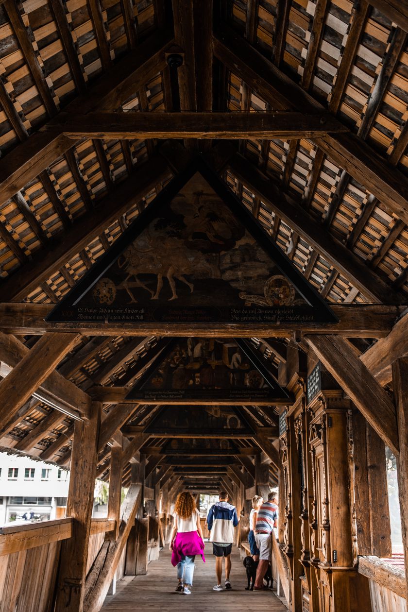 Inneres der Spreuerbrücke in Luzern: Mehrere Personen gehen durch den überdachten Holzgang, über ihnen sind dreieckige Giebelbilder mit Totentanz-Motiven zu sehen.