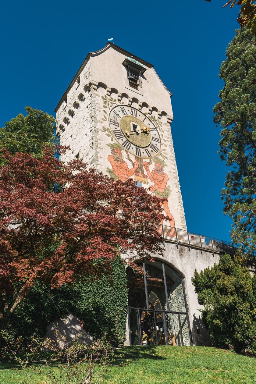 Der Zytturm der Museggmauer in Luzern, von unten fotografiert. An der Turmfassade ist eine grosse Uhr mit römischen Zahlen und ein farbiges Fresko mit figürlichen Darstellungen zu sehen. Der Turm ist von Bäumen und einer Grünfläche umgeben.