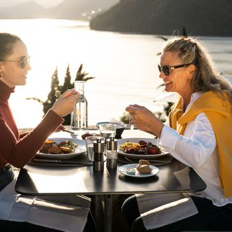 Zwei Frauen sitzen sich an einem gedeckten Aussentisch gegenüber und essen gemeinsam. Auf dem Tisch stehen Teller mit Essen, Gläser, Salz- und Pfefferstreuer sowie eine Wasserflasche. Im Hintergrund ist ein helles Gewässer mit Hügeln zu sehen.