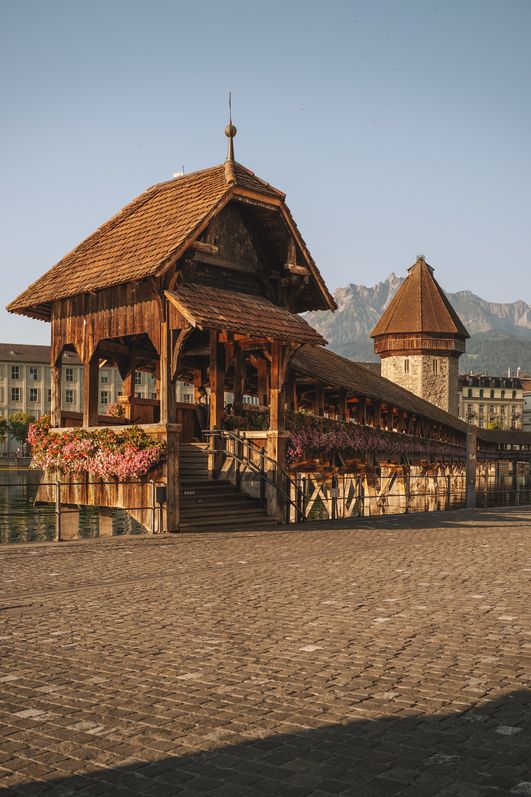 Holzbrücke mit überdachtem Pavillon und Blumenschmuck am Wasser, im Hintergrund ein Turm, Stadtgebäude und Berge unter klarem Himmel.