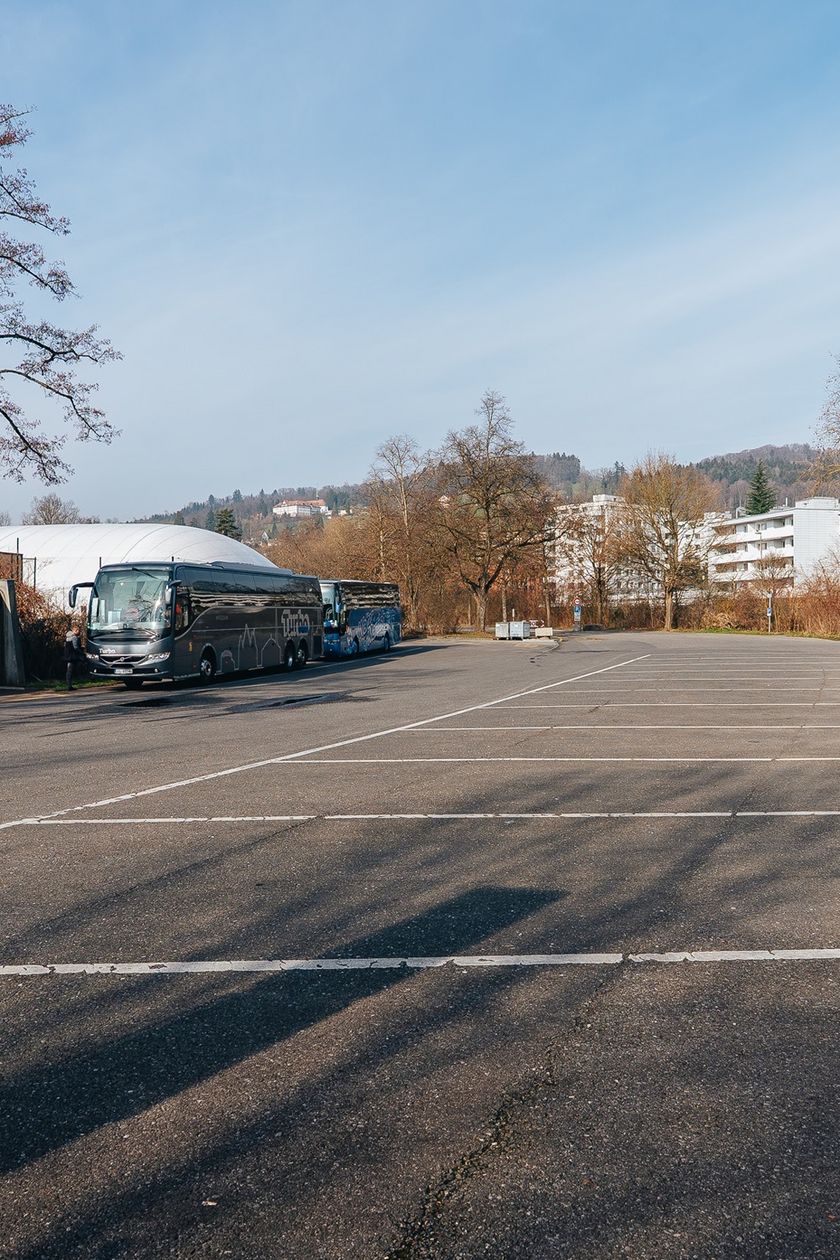 Weitläufiger Busparkplatz beim Lido Luzern (Brüel) mit zwei geparkten Reisecars, kahlen Bäumen und Wohnhäusern im Hintergrund, aufgenommen bei bewölktem Winterhimmel.