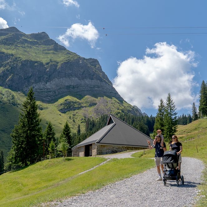 Eine Familie mit Kinderwagen geht auf einem Kiesweg durch eine alpine Landschaft mit Wiesen, Tannen und einem Bergmassiv; im Hintergrund stehen eine kleine Hütte und Seilbahnkabel am Himmel.
