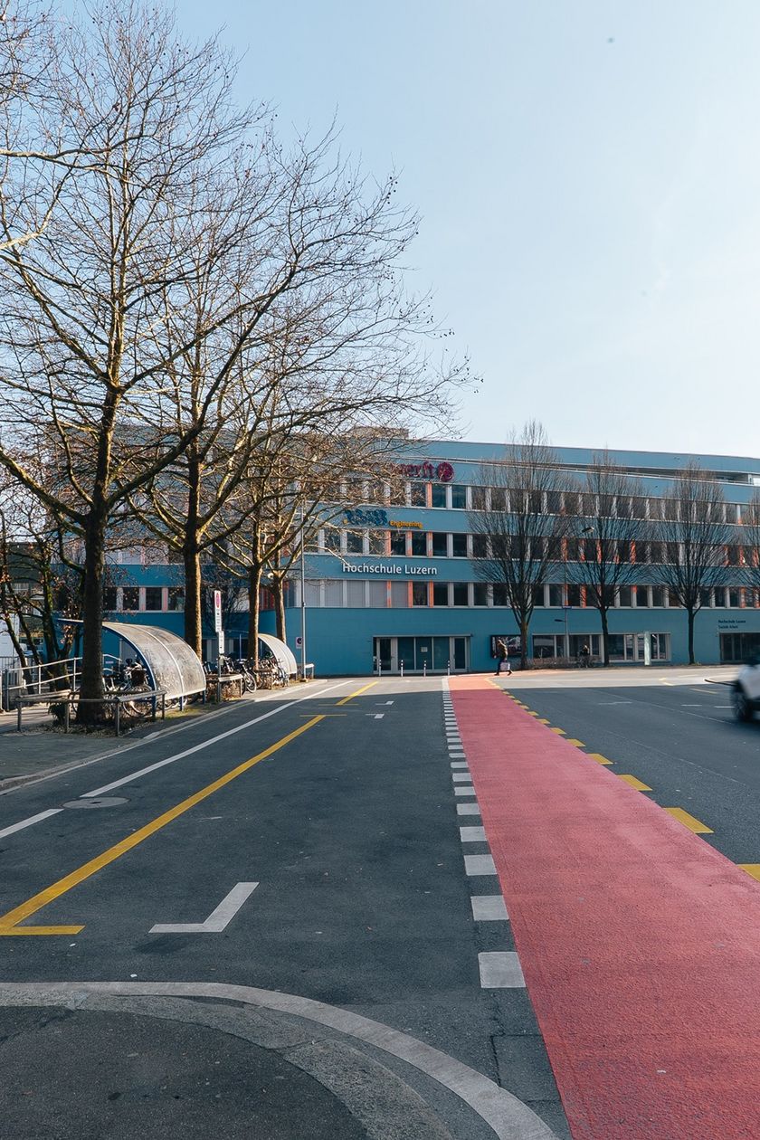 Strassenansicht der Haltekante am Inseliquai in Luzern mit rot markiertem Haltebereich, einem fahrenden Auto, kahlen Bäumen sowie dem Gebäude der Hochschule Luzern im Hintergrund.