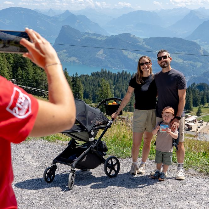 Eine Person im roten Shirt fotografiert eine Familie mit Kind neben einem Kinderwagen auf einem Bergweg; im Hintergrund sind Alpen, ein See und eine Bergwiese zu sehen.