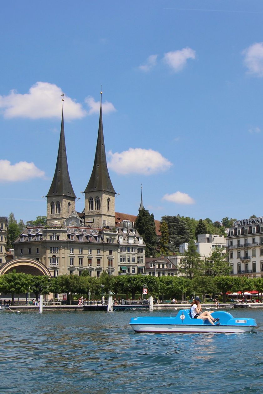 Eine Person auf einem blauen Pedalo auf dem Vierwaldstättersee, im Hintergrund die Doppeltürme der Hofkirche St. Leodegar und das Hotel National an der Luzerner Uferpromenade.