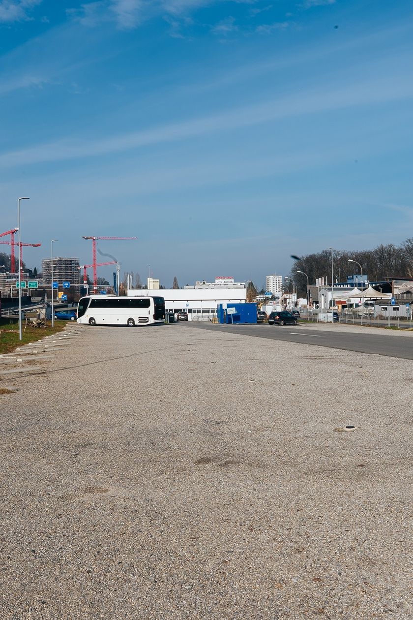 Grosser, weitgehend leerer Schotterplatz in einem Gewerbegebiet in Kriens. Ein weisser Reisecar steht im mittleren Hintergrund. Links eine bemalte Wand, rechts Gewerbegebäude. Im Hintergrund Baukräne und bewaldete Hügel unter blauem Himmel.