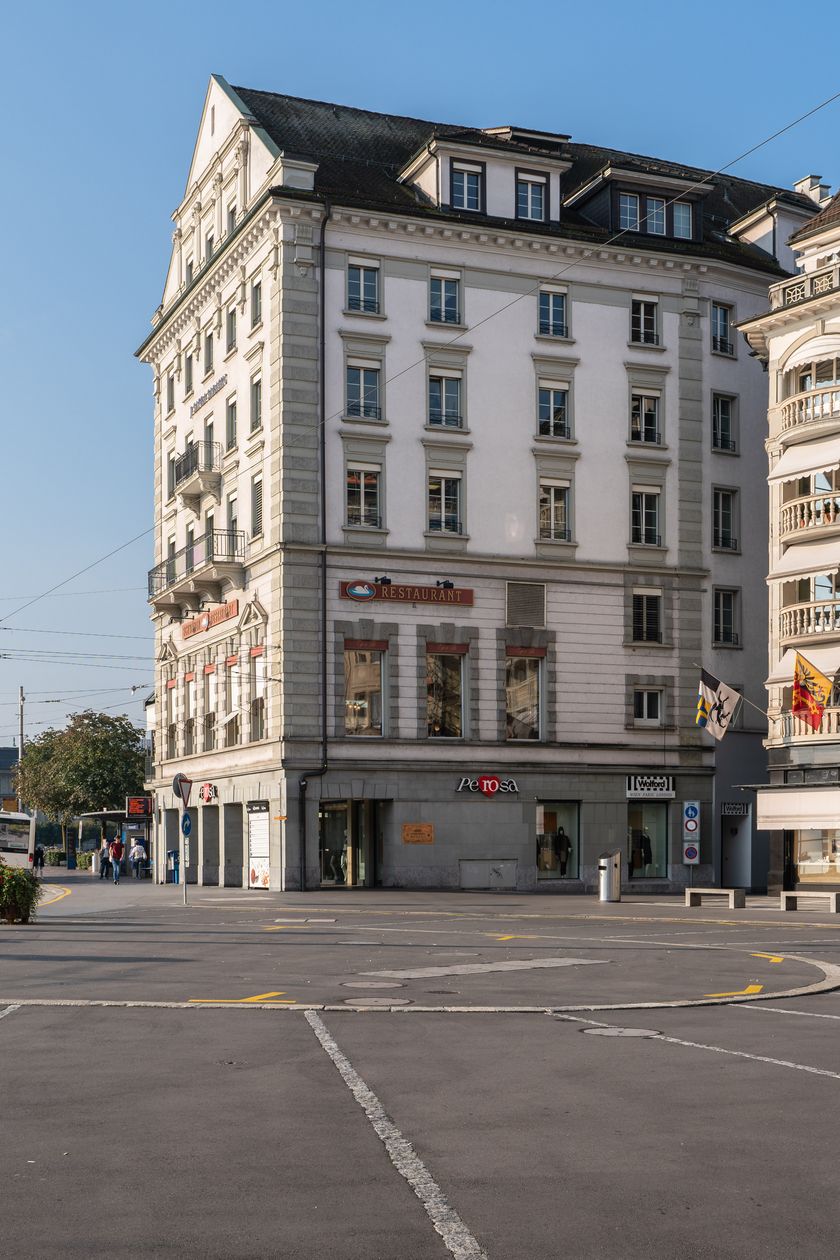 Der Schwanenplatz in Luzern bei sonnigem Wetter: eine grosse, weitgehend leere Platzfläche, flankiert von historischen Stadtgebäuden mit den Fassaden der Uhrengeschäfte Bucherer und Rolex. Links im Bild rote Blumensäulen und ein Bus, im Hintergrund weitere Stadtgebäude und der Luzerner Bahnhof.