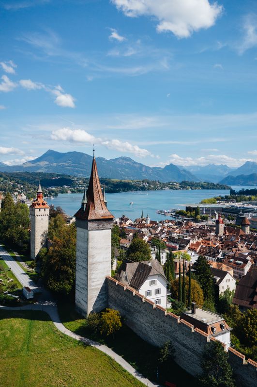 Luftaufnahme von Luzern mit mittelalterlichen Türmen, Stadtmauer, dicht bebauter Altstadt und dem Vierwaldstättersee im Hintergrund; dahinter Berge unter blauem Himmel mit einzelnen Wolken.