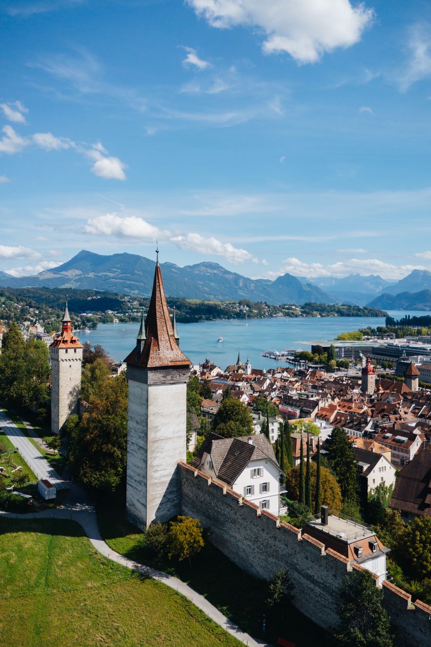 Luftaufnahme von Luzern mit mittelalterlichen Türmen, Stadtmauer, dicht bebauter Altstadt und dem Vierwaldstättersee im Hintergrund; dahinter Berge unter blauem Himmel mit einzelnen Wolken.