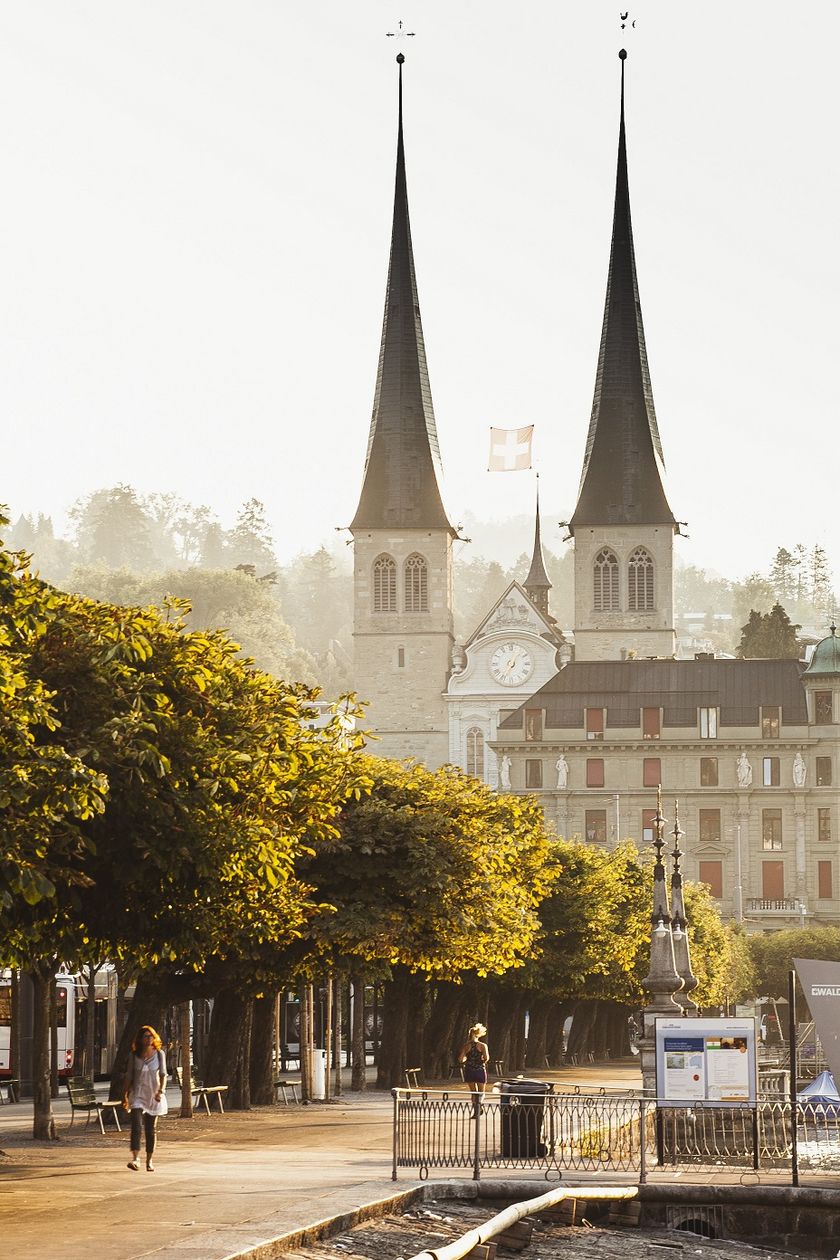 Zwei spitze Kirchtürme der Hofkirche St. Leodegar ragen über Baumkronen und historische Gebäude hinaus. Im Vordergrund flanieren zwei Frauen entlang einer Uferpromenade.
