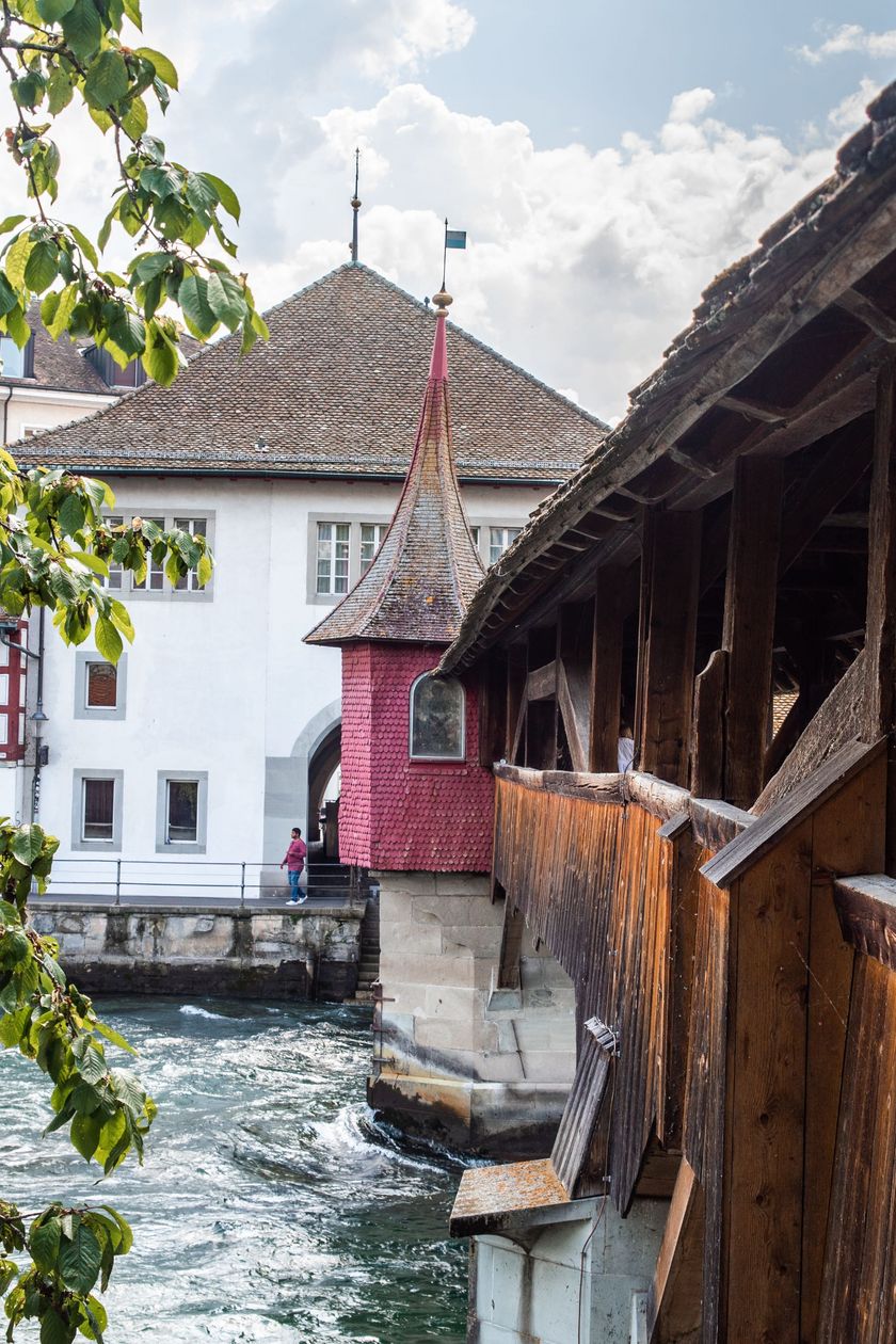 Blick von der überdachten Spreuerbrücke über die Reuss in Luzern. Im Vordergrund die hölzerne Brückenkonstruktion, in der Bildmitte ein roter Treppenturm mit Spitzdach, im Hintergrund historische Altstadtgebäude und ein Laubbaum.