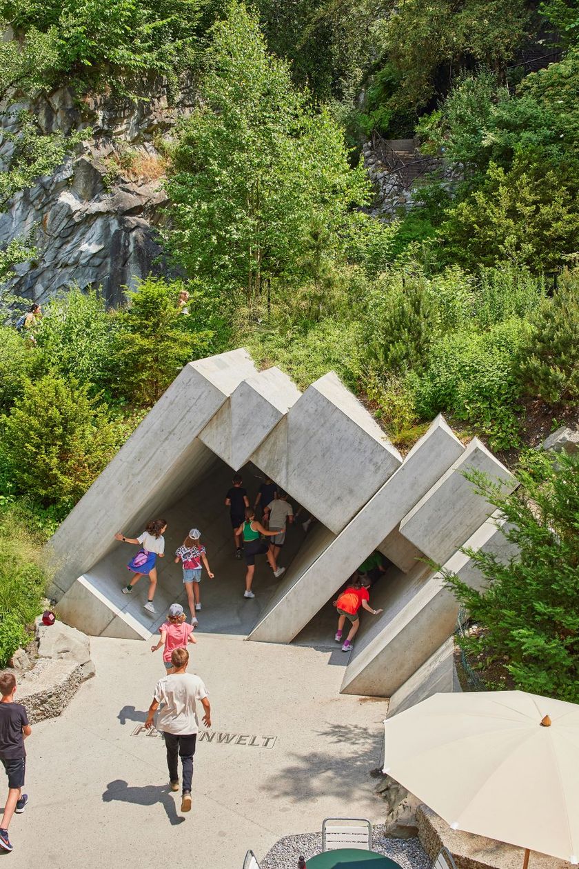 Vogelperspektive auf den geometrisch geformten Betoneingang zur Felsenwelt im Gletschergarten Luzern. Mehrere Kinder und Erwachsene gehen in den Eingang hinein, umgeben von grüner Vegetation und Felswänden.