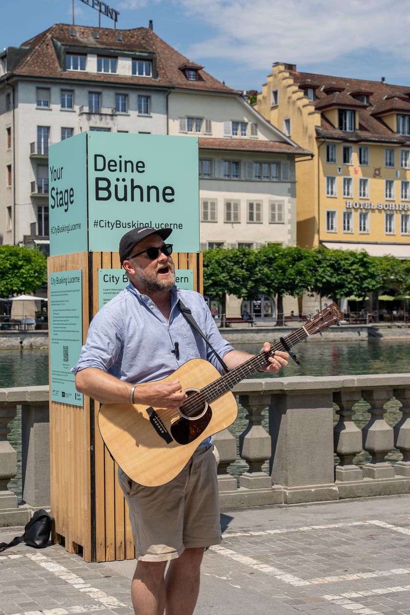 Ein Mann mit Sonnenbrille und Kappe spielt auf einer Gitarre vor einer hellblauen Box mit der Aufschrift «Deine Bühne» und «Your Stage». Im Hintergrund stehen Häuser am Ufer mit der Aufschrift «Hotel Schiff».