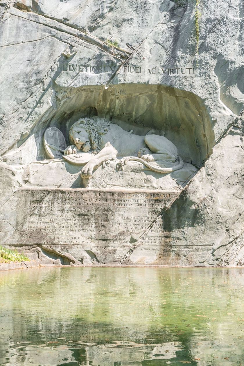 In eine graue Felswand eingehauenes Relief eines Löwen, umgeben von lateinischen Inschriften. Im Vordergrund ein stiller Teich, der die Szenerie widerspiegelt. Grüne Vegetation rahmt das Denkmal seitlich ein.