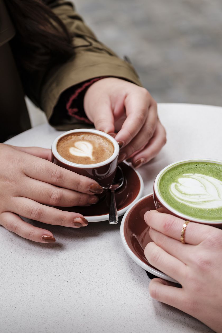 Zwei Personen halten Cappuccino und Matcha Latte mit Herzmuster an einem kleinen Tisch.