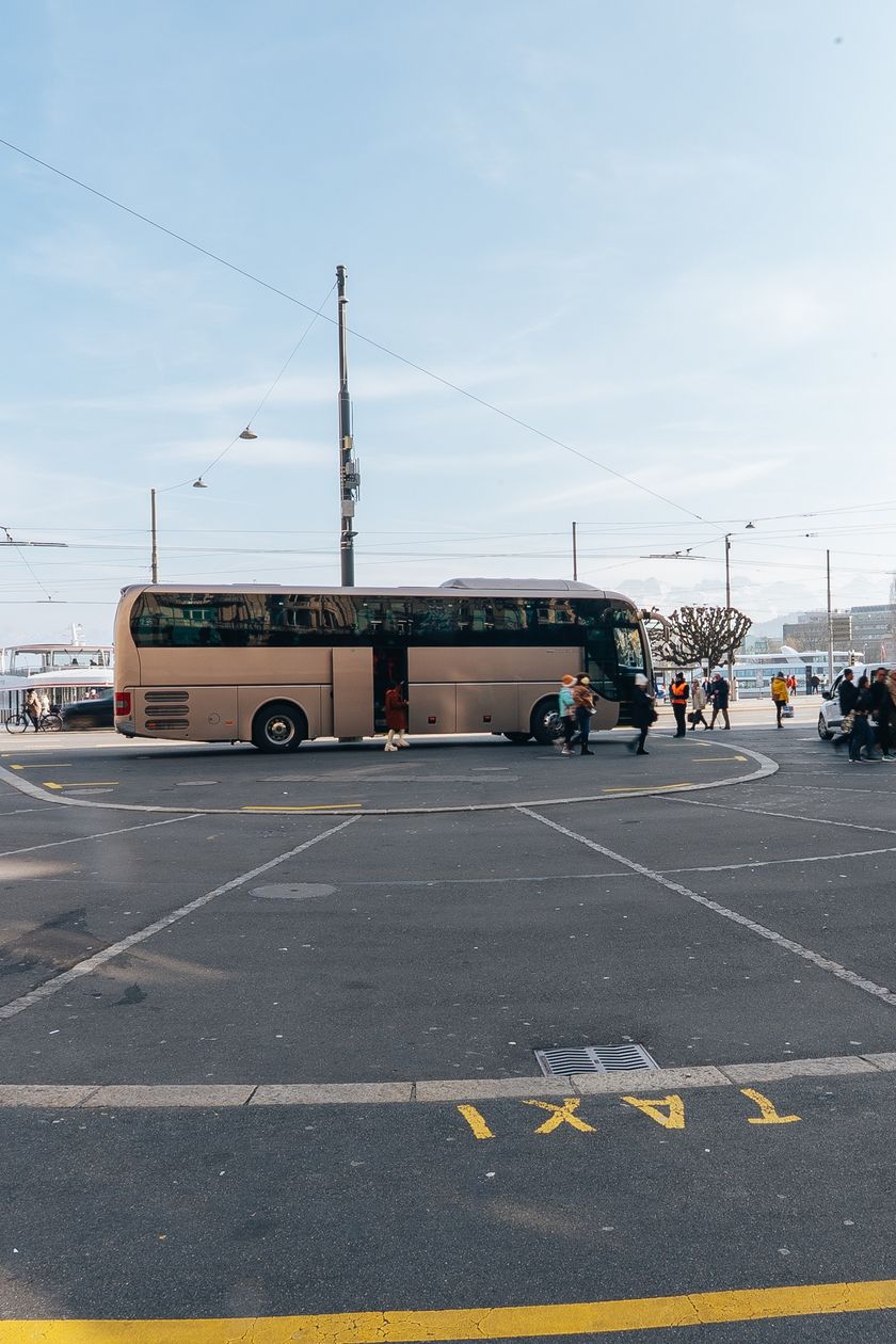 Ein goldbeiger Reisecar steht auf dem Schwanenplatz in Luzern. Im Vordergrund ist eine Taxi-Markierung auf dem Asphalt zu sehen, im Hintergrund der Vierwaldstättersee und ein Kursschiff. Mehrere Fussgänger überqueren den Platz.