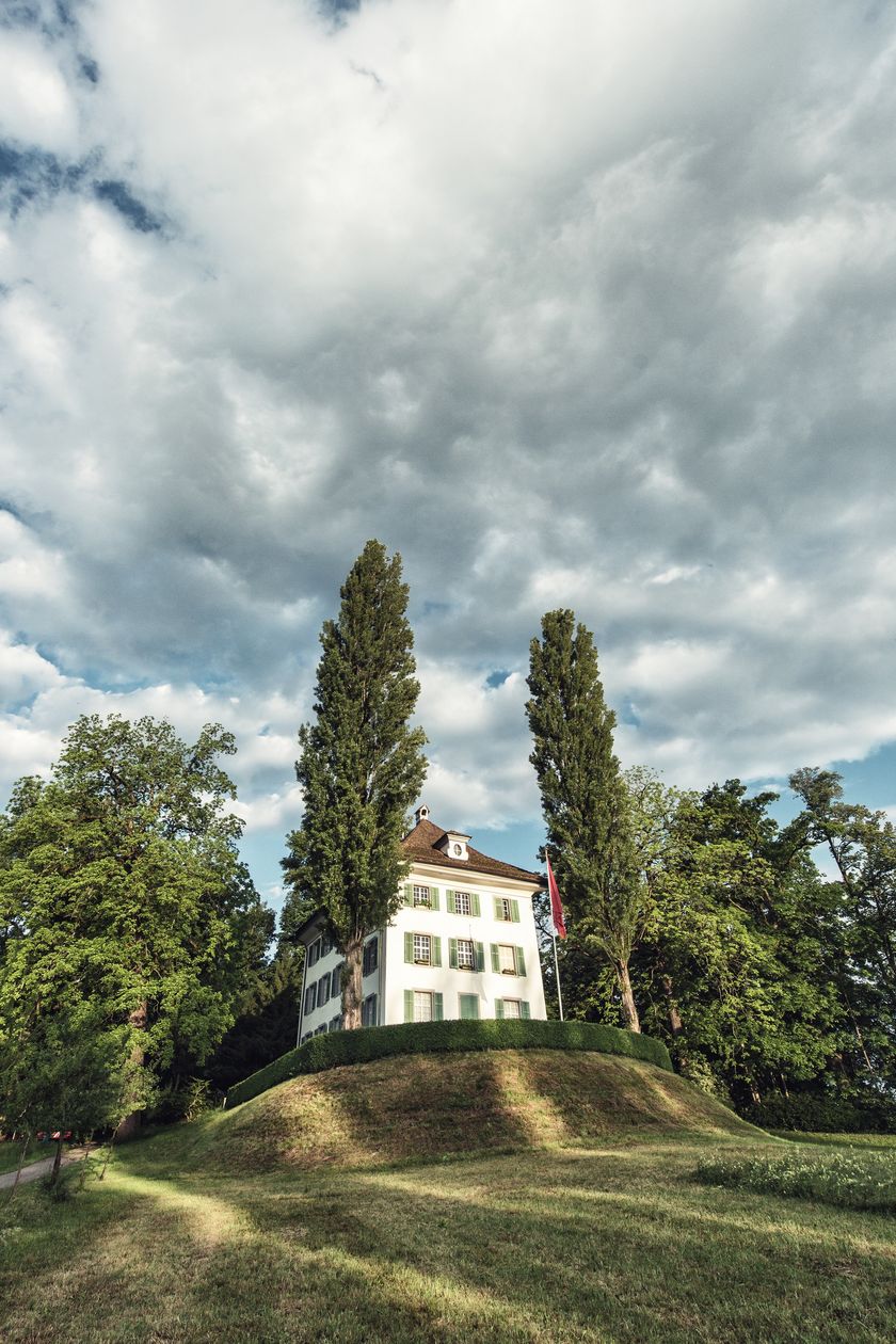 Weisses zweigeschossiges Herrenhaus mit grünen Fensterläden auf einem Grashügel, flankiert von zwei hohen Pappeln, vor bewölktem Himmel – Villa Tribschen, Sitz des Richard Wagner Museums in Luzern.