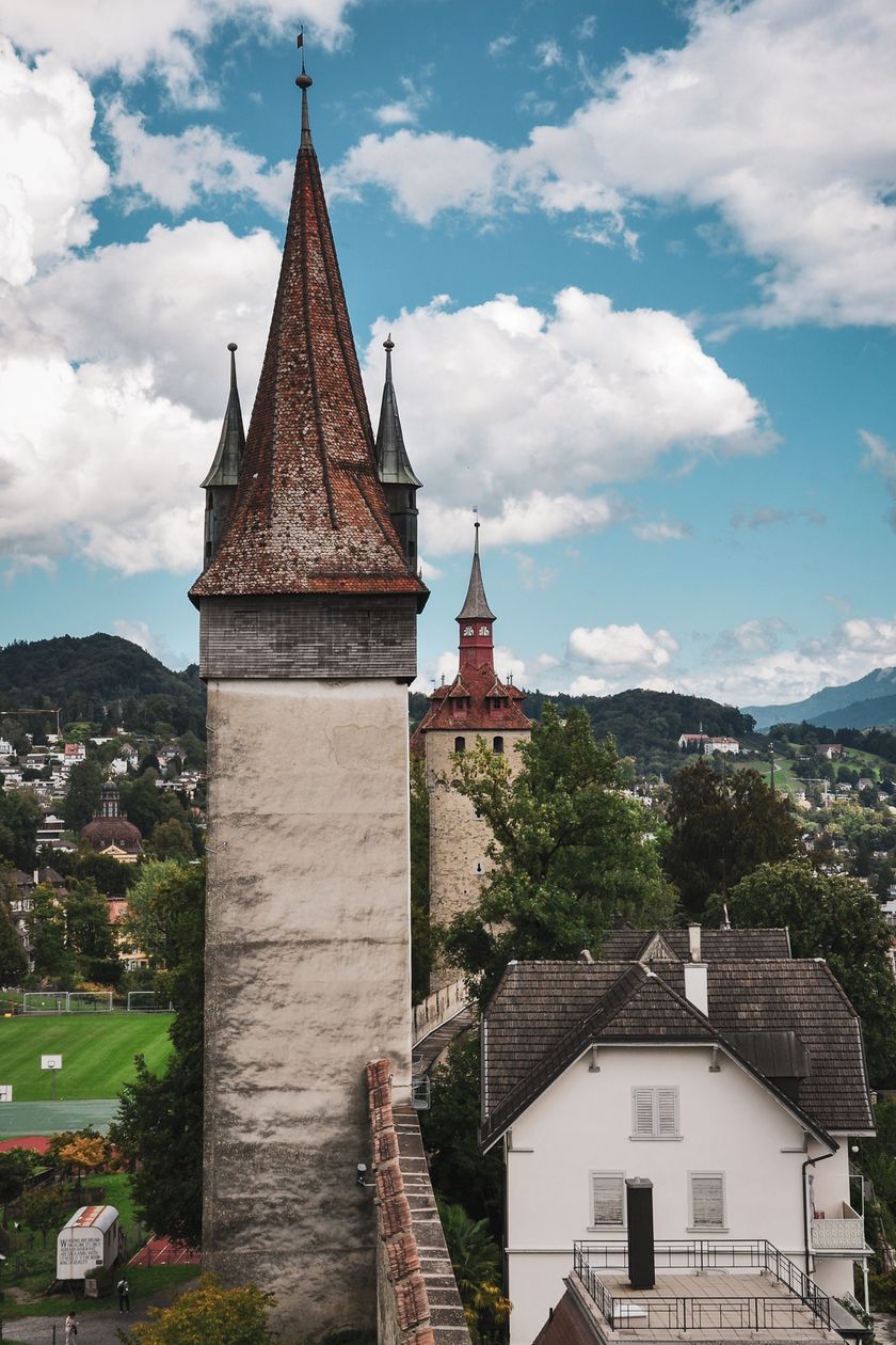 Zwei Türme der Museggmauer in Luzern ragen vor einem bewölkten Himmel auf. Im Vordergrund ein schlanker Turm mit spitzem Ziegeldach, dahinter ein weiterer Turm mit rotem Helm. Unten sind Wohnhäuser und eine Sportanlage sichtbar.